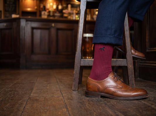 Burgundy Louluu Galloper ribbed sock between a turned-up navy trouser hem and a brown brogue on a wooden pub floor