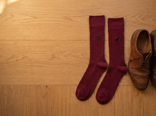 A pair of burgundy Galloper Ribbed socks with a small embroidered horse, laid on a wooden floor next to brown brogues in soft autumn light.