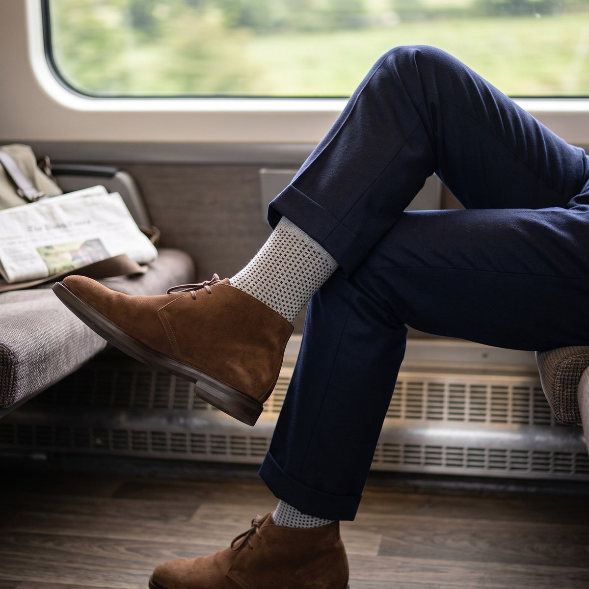 Man on a train by the window wearing grey birdseye lambswool socks with navy trousers and suede chukka boots