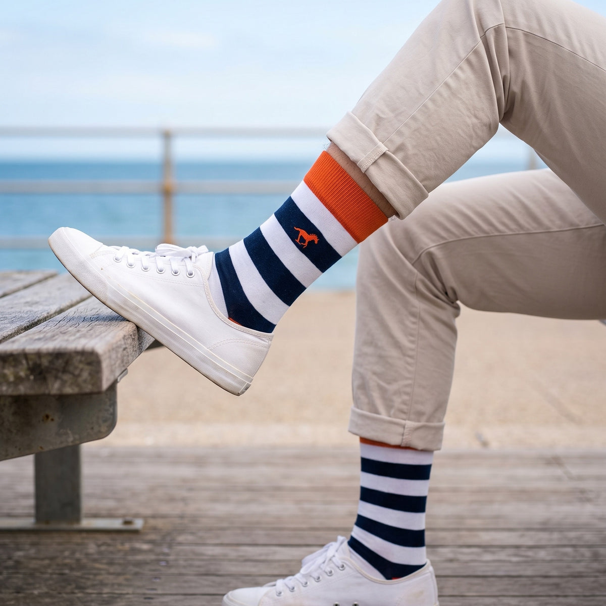 Man on a seaside bench wearing navy and white Galloper Rugby stripe socks with beige chinos and white canvas sneakers