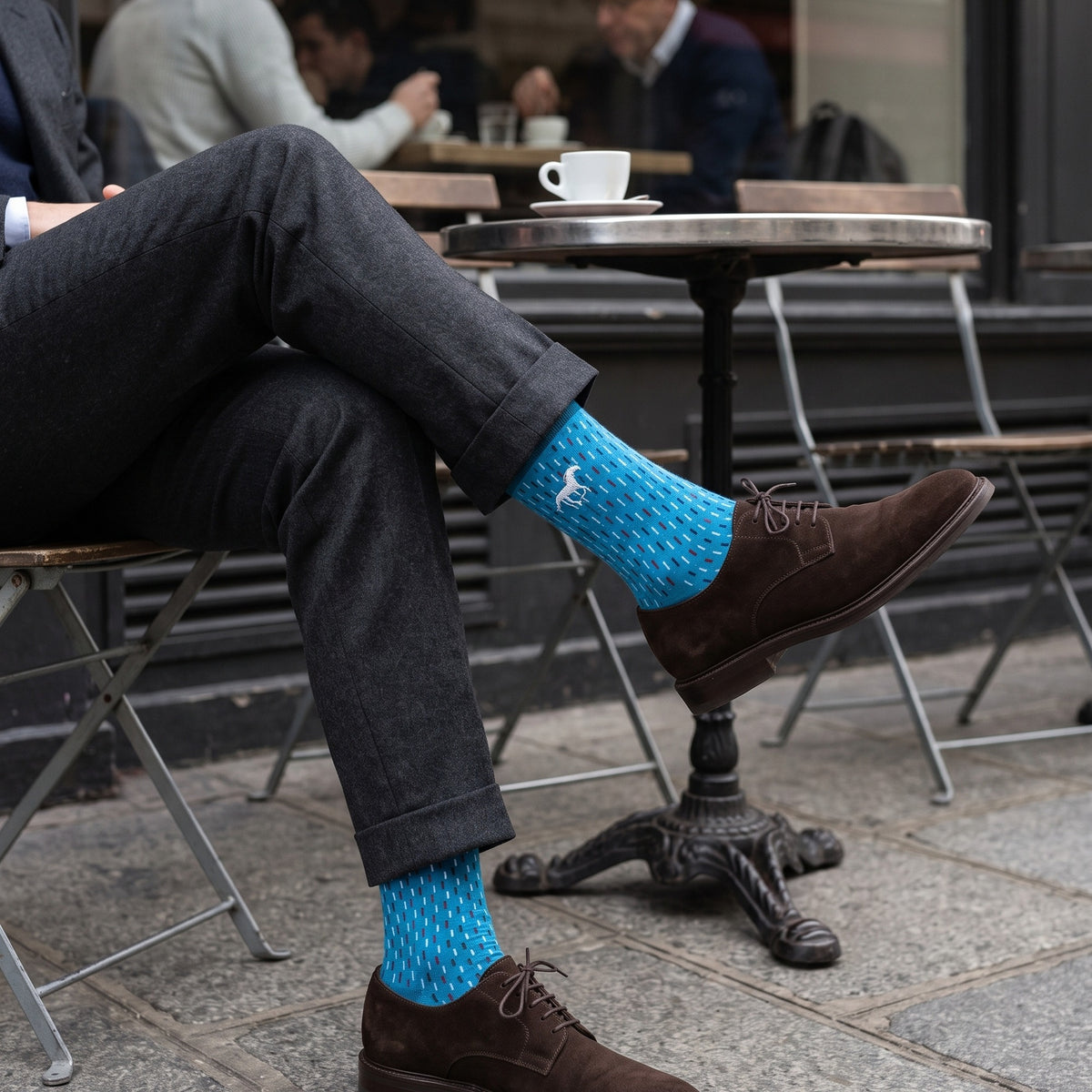 Man at a Paris café wearing blue Galloper Stitch socks with white horse emblem, grey trousers and suede derby shoes