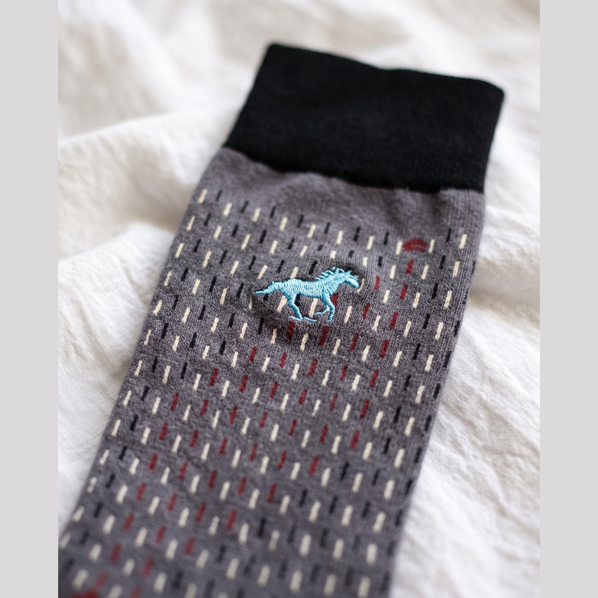 Close-up of grey Galloper Stitch sock with black cuff, dash stitch pattern and light blue embroidered horse