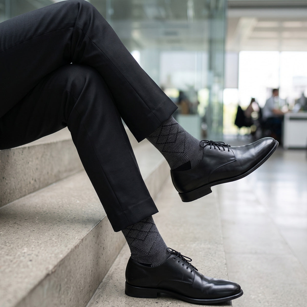 Man on concrete steps in an office atrium wearing pinstripe diamond socks with black trousers and derby shoes
